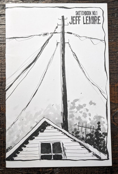 A sketchbook featuring a black and white hand-drawn illustration of a house roof, power lines, and a utility pole, with the text 'SKETCHBOOK NO.1 JEFF LEMIRE' in the top right corner, placed on a dark wooden surface.
