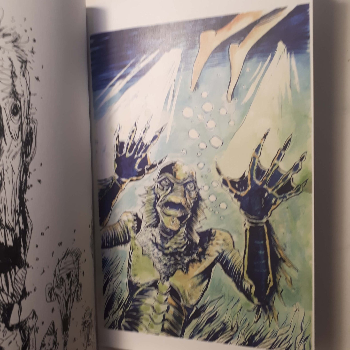A close-up of an illustrated page showing a monstrous creature with clawed hands reaching upward in a blue-green underwater scene, with partial black line art visible on the left page.