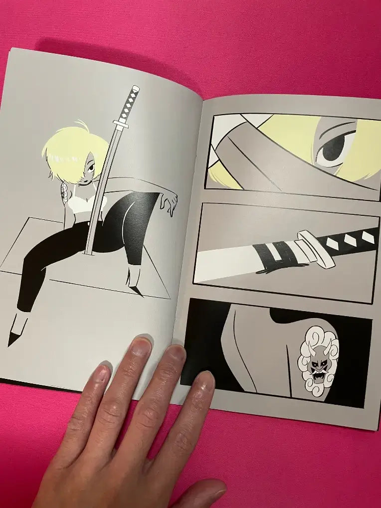 An open manga or comic book showing a black-and-white illustrated page with a blonde character holding a sword, alongside close-up panels, photographed on a bright pink background with a hand holding the page.