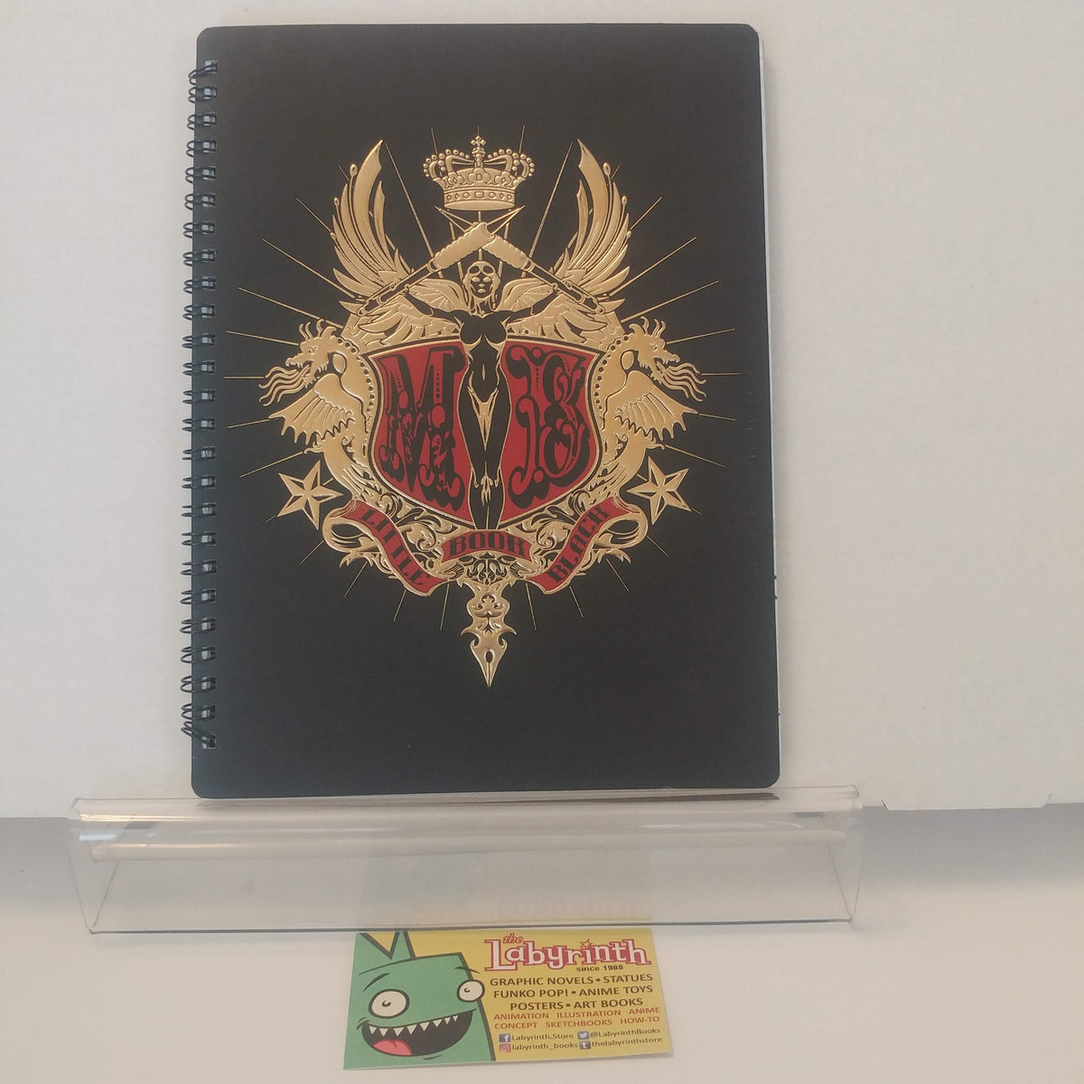 A black spiral-bound notebook featuring a large gold and red crest design on the cover, displayed on a clear acrylic stand against a white background.