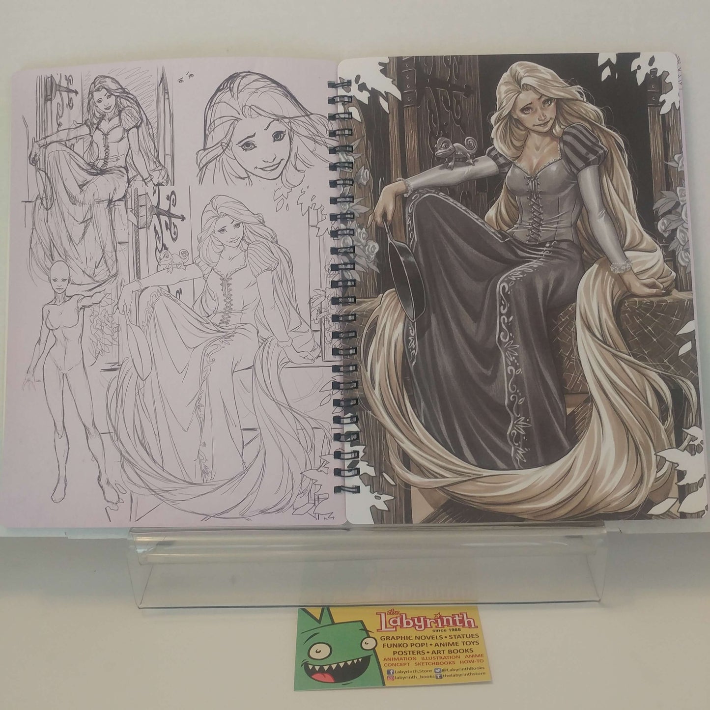 An open spiral-bound sketchbook or art print showing pencil line drawings of a long-haired female character on the left and a fully colored illustration on the right, displayed on a clear stand with a small card from thelabyrinthstore below.