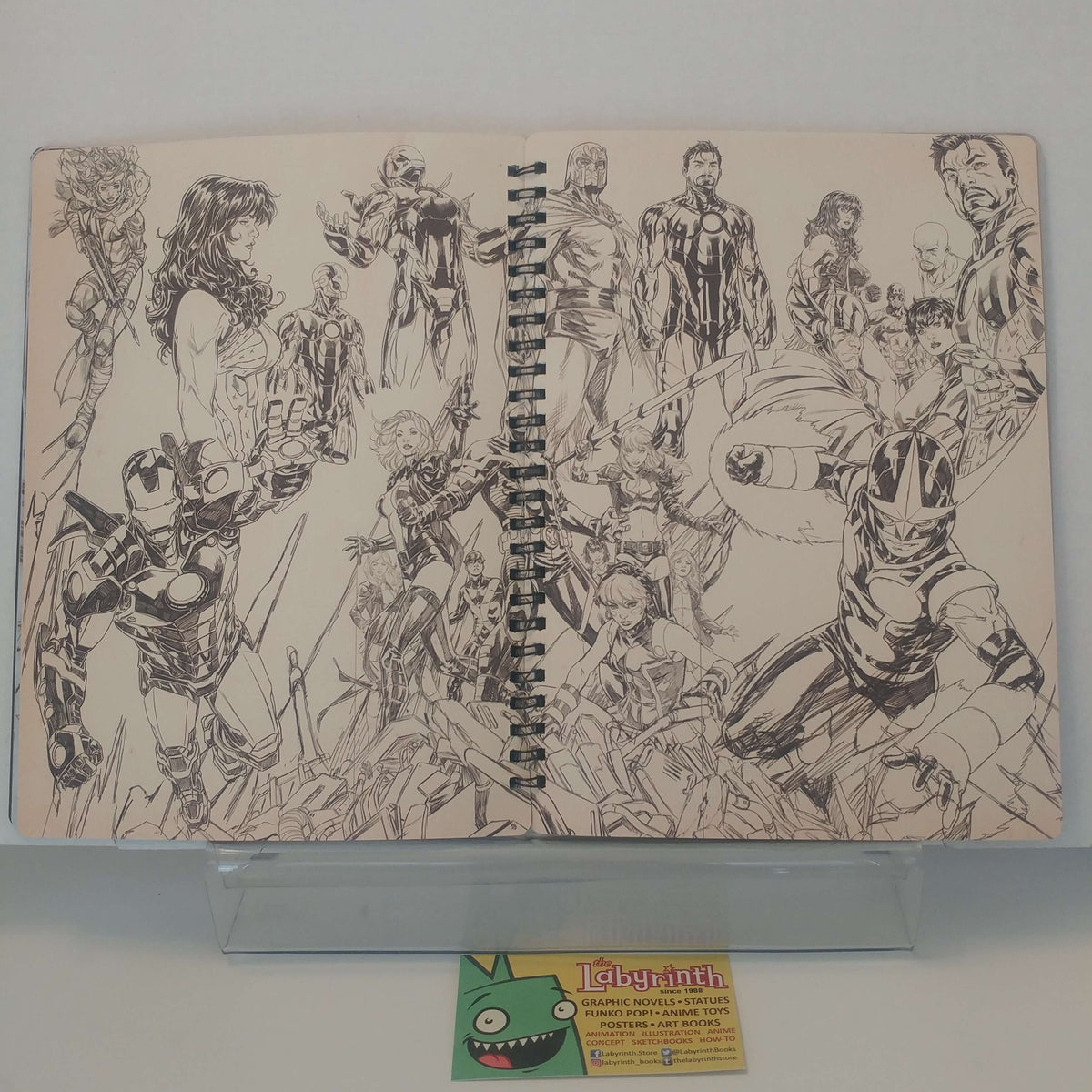 A spiral-bound sketchbook featuring detailed black ink illustrations of Marvel superheroes on off-white paper, displayed on a clear stand with a small card from thelabyrinthstore in front.