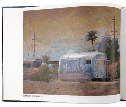 An open art book or magazine showing a painted scene of a trailer-style home in a beach-like setting, with utility poles, a palm tree, and a dusty sky. The page has white margins and small text at the bottom reading 'BOMBAY BEACH #2 24632'.