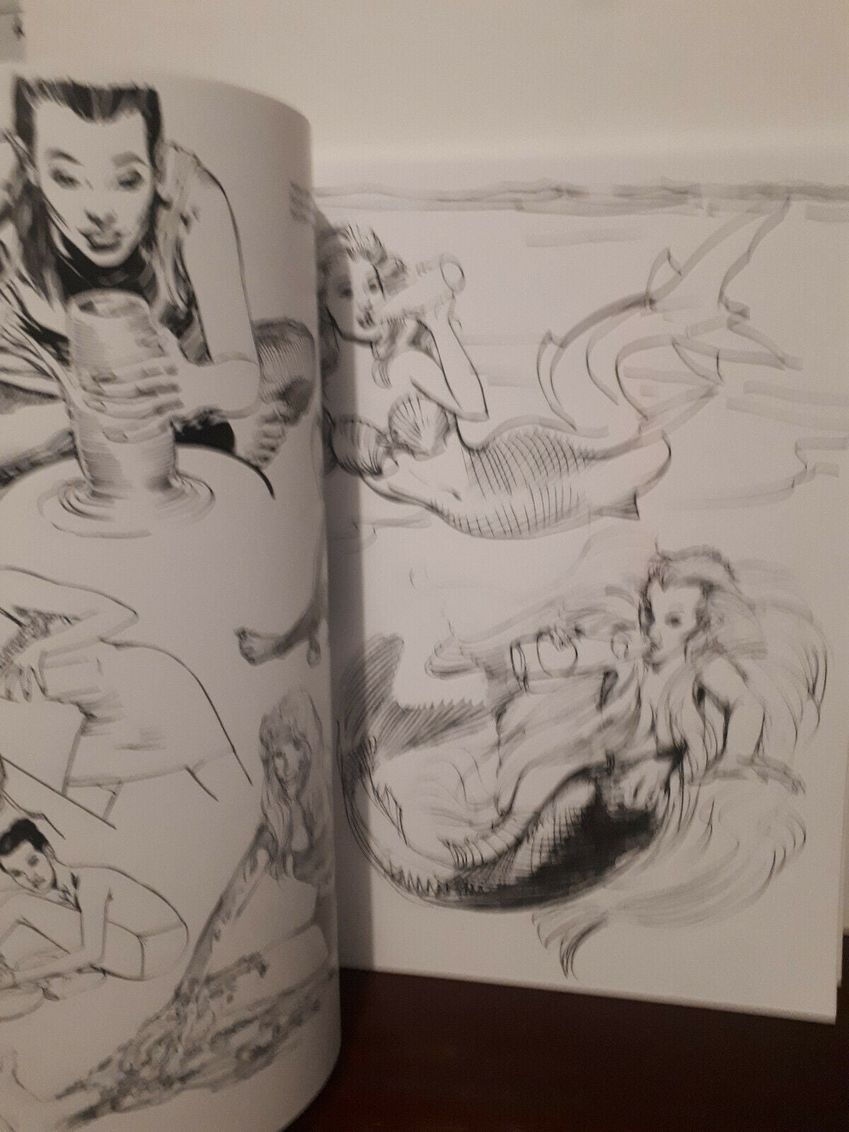 An open sketchbook or art book showing black ink mermaid and figure studies on white pages, photographed indoors on a dark surface against a light wall.