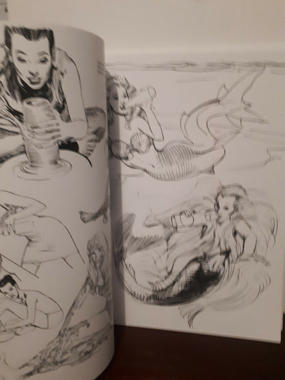 An open sketchbook or art book showing black ink mermaid and figure studies on white pages, photographed indoors on a dark surface against a light wall.