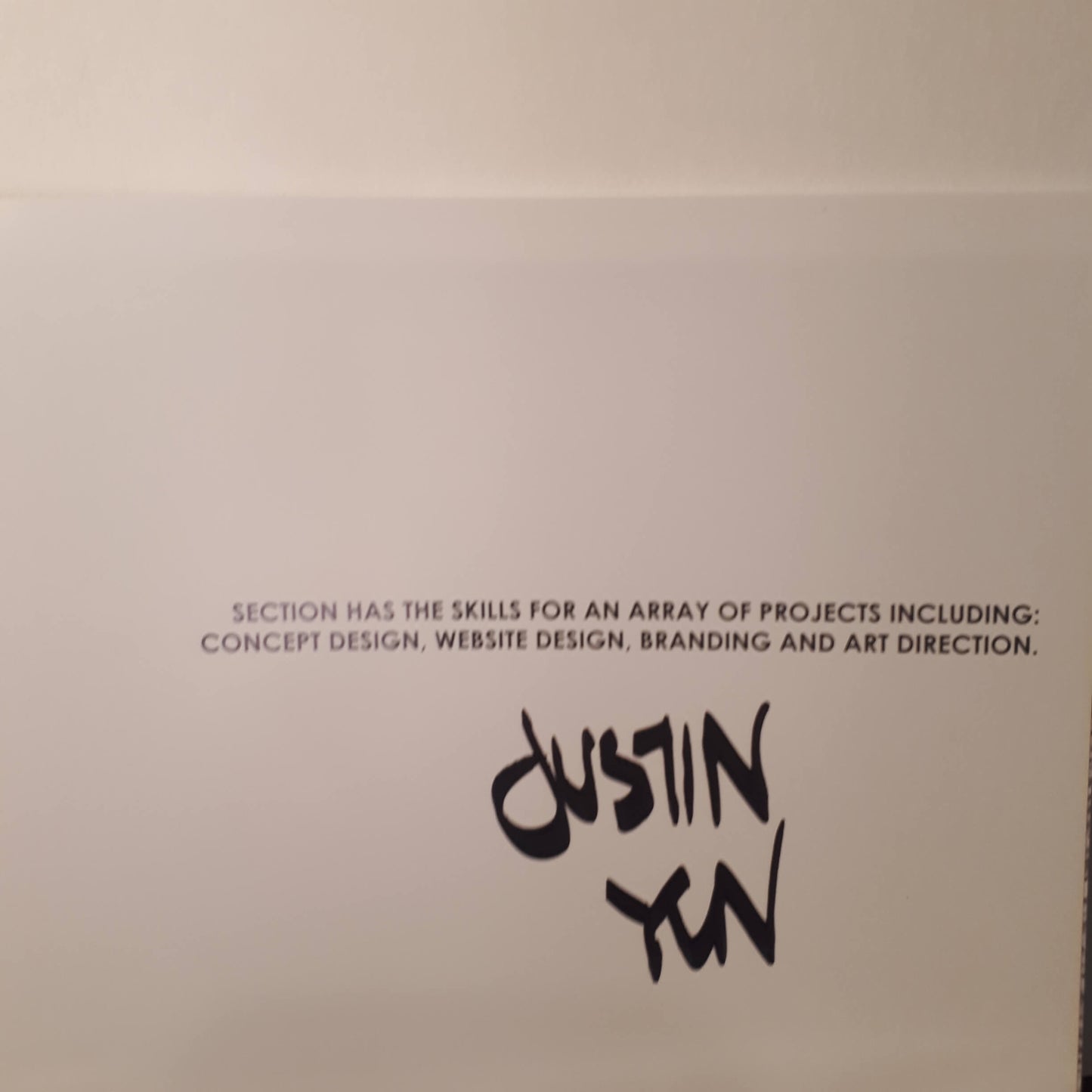 A white card or paper featuring black printed text that reads, 'SECTION HAS THE SKILLS FOR AN ARRAY OF PROJECTS INCLUDING: CONCEPT DESIGN, WEBSITE DESIGN, BRANDING AND ART DIRECTION.' Below this text is the name 'Dustin Yan' written in a stylized black script.
