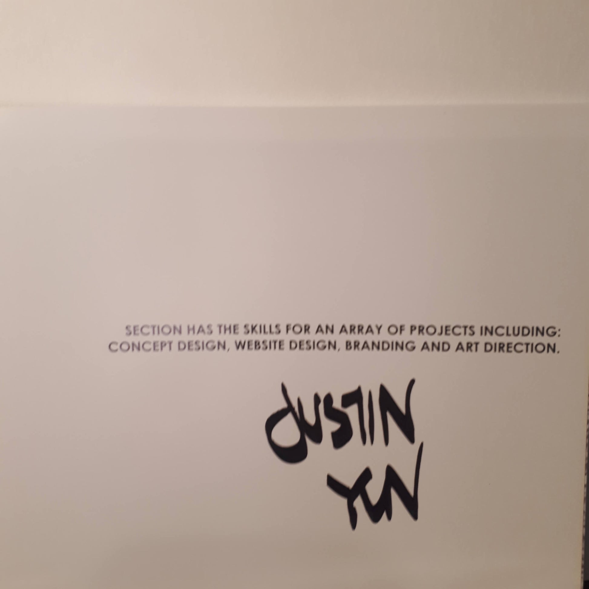 A white card or paper featuring black printed text that reads, 'SECTION HAS THE SKILLS FOR AN ARRAY OF PROJECTS INCLUDING: CONCEPT DESIGN, WEBSITE DESIGN, BRANDING AND ART DIRECTION.' Below this text is the name 'Dustin Yan' written in a stylized black script.