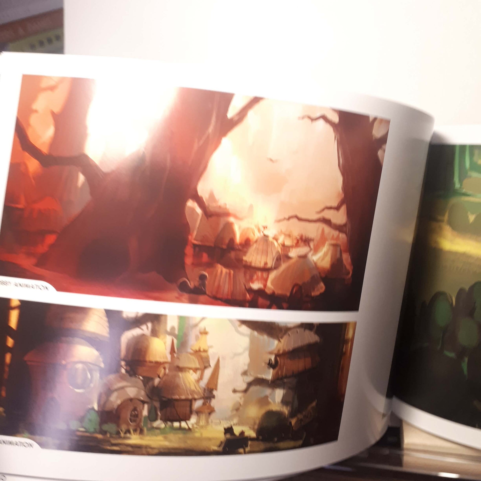 An open printed art book or magazine showing colorful fantasy landscape illustrations on the pages, photographed indoors with some glare and partial surrounding objects visible.