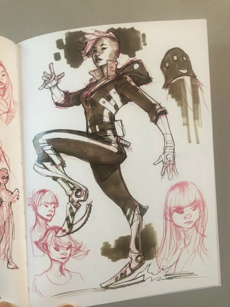 An open sketchbook showing concept art illustrations of a stylized female character in a dark outfit, with pink hair and smaller character sketches around her on the page.