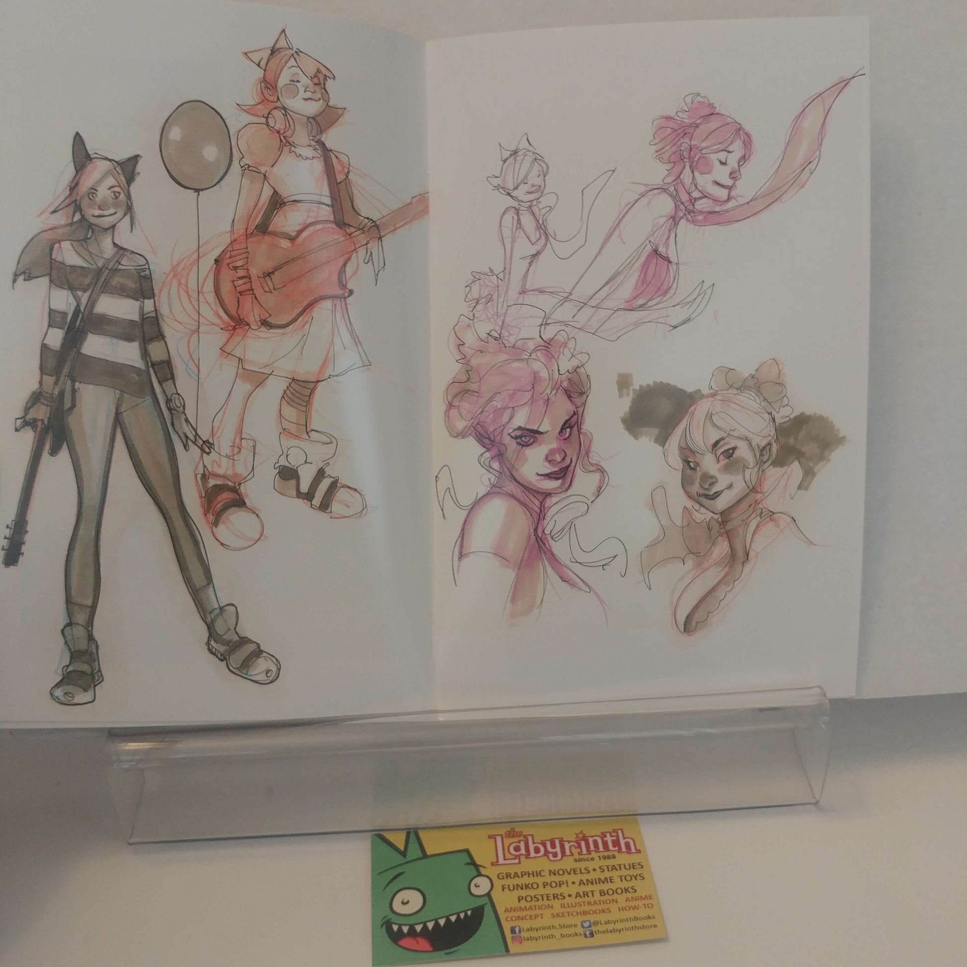 An open sketchbook displaying character concept art, with two illustrated cartoon figures and several pink-toned character studies on white pages. A small card at the bottom reads 'Labyrinth'.