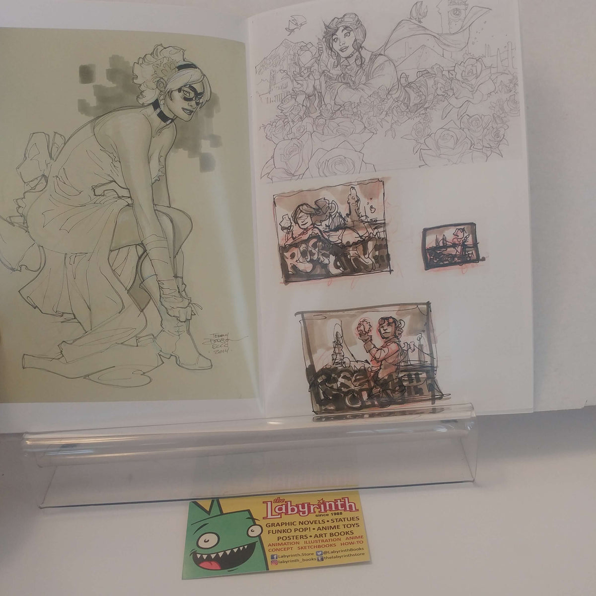 An open sketchbook or art portfolio displayed on a clear stand, showing line drawings and small painted scenes. A small card at the bottom reads 'labyrinth' and appears to be part of the presentation.