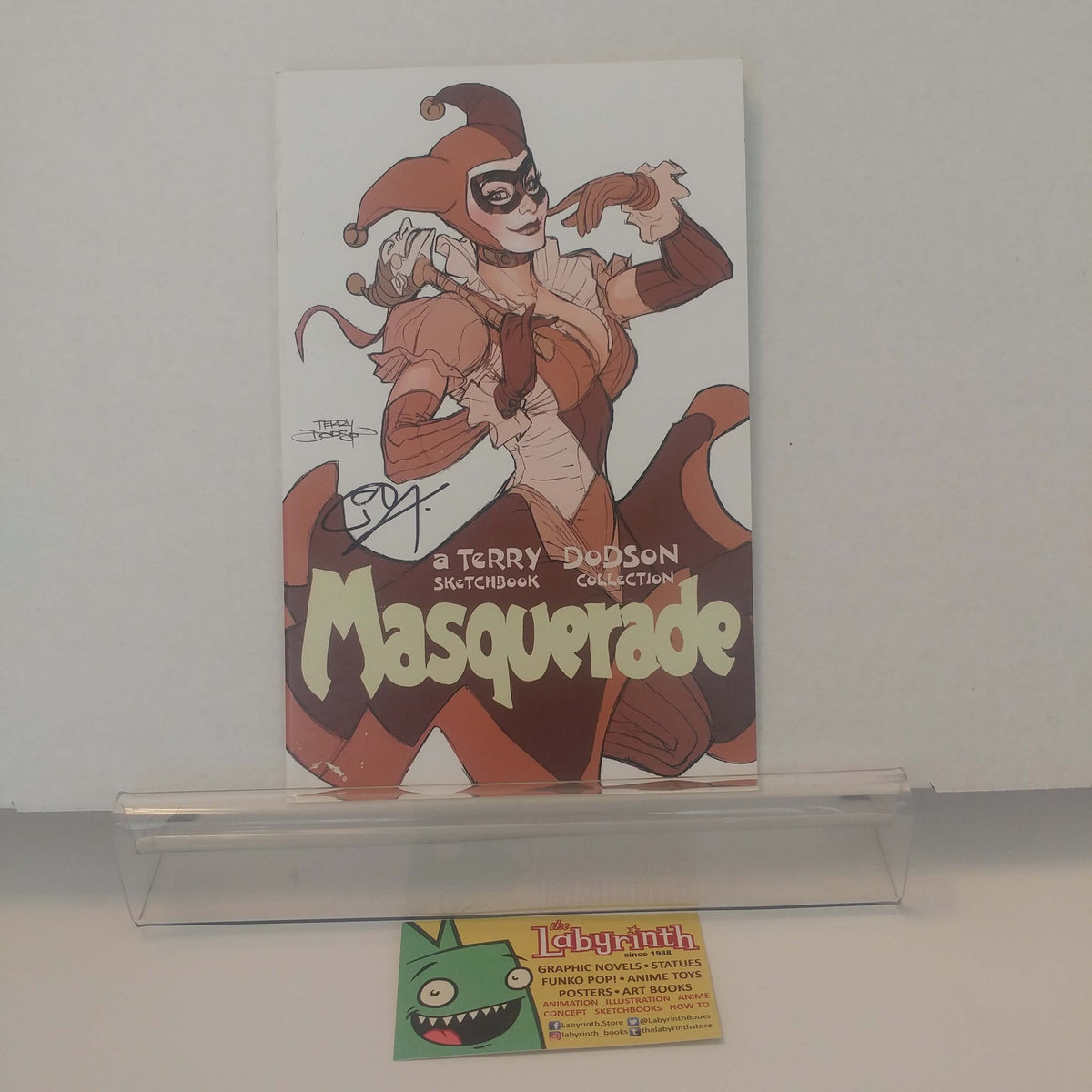 A signed comic book titled 'Masquerade' by Terry Dodson, featuring an illustrated character in a jester costume, displayed on a clear acrylic stand with a Labyrinth card below it.