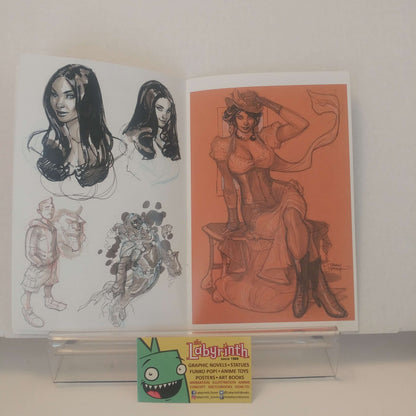 An open sketchbook or art portfolio displayed on a clear acrylic stand, showing character illustrations. The left page has black-and-white and gray sketches, and the right page has a colored pencil drawing of a seated woman on an orange paper background.