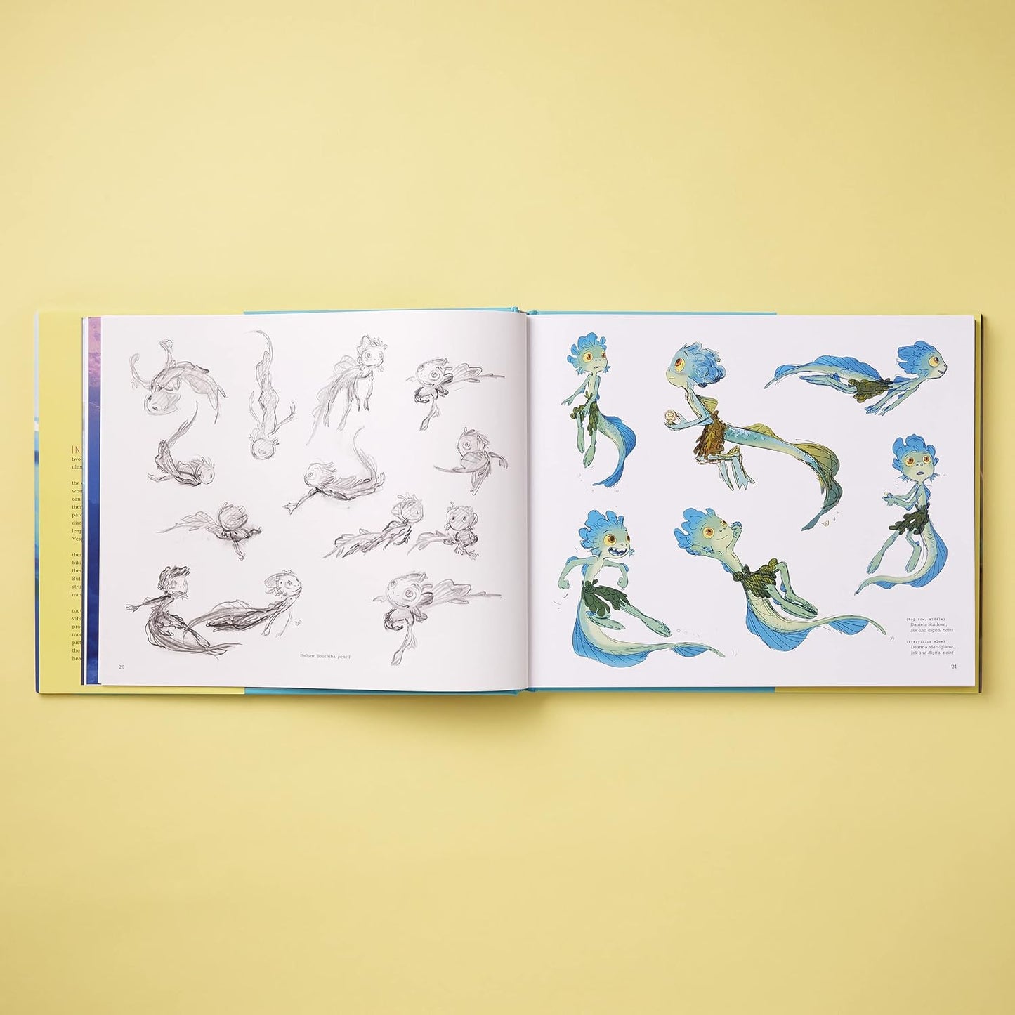 An open art book or concept sketchbook laid flat on a pale yellow background, showing character designs and pencil studies on both pages. The illustrations feature blue, green, and brown fantasy figures on white paper.
